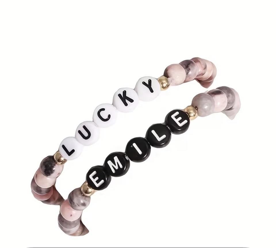 Close-up of a finished two-piece affirmation bracelet set: one bracelet with white letter beads spelling LUCKY and one with black letter beads spelling EMILE, both with pink and gray stone beads and gold spacers