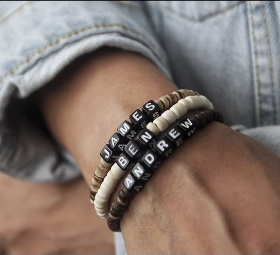 Wrist wearing stacked personalized name bracelets with letter beads spelling James, Ben, and Andrew on wood-tone beads
